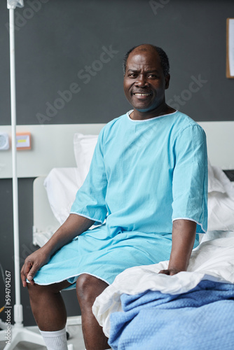 Vertical image of African American patient in shirt smiling at camera while sitting on bed in hospital ward