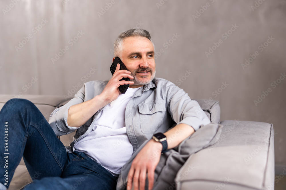 Successful and smiling middle-aged man in white shirt talking by the phone at home. Sitting on the couch and looking in front of him. Successful and smiling middle-aged man in white shirt talking by the phone at home. Sitting on the couch and looking in front of him.