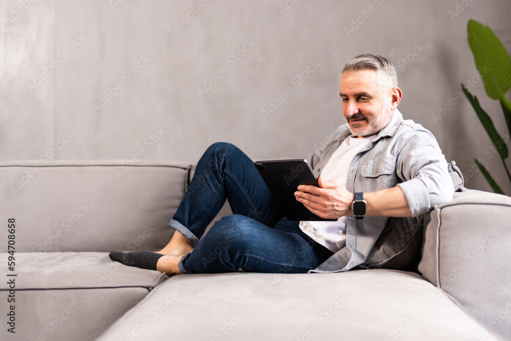 Portrait of a serious handsome bearded guy, young middle aged man is using his tablet laptop, relaxing, lying at home on sofa couch, looking at screen, reading electronic book