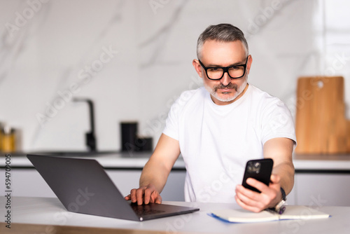 Handsome mature man talking on mobile phone while using laptop at the table at home