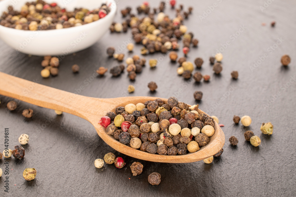 A mixture of four peppercorns on a white saucer with a wooden spoon, macro, on slate stone. A mixture of four peppercorns on a white saucer with a wooden spoon, macro, on slate stone.