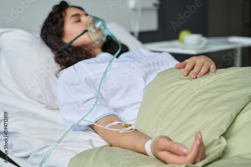 Young patient with catheter in her arm and oxygen mask lying on bed in hospital ward