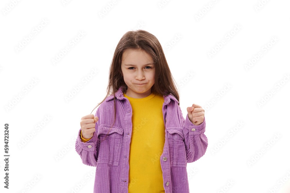 Selective focus on a Caucasian curious angry little child girl clenching fists, expressing negative emotions - anger and dissatisfaction, looking at camera, isolated on white background. Copy ad space Selective focus on a Caucasian curious angry little child girl clenching fists, expressing negative emotions - anger and dissatisfaction, looking at camera, isolated on white background. Copy ad space