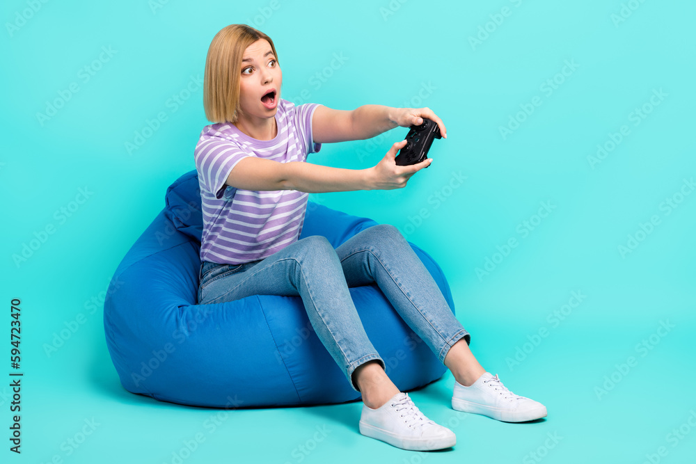 Full length photo of ecstatic girl stylish t-shirt play excited game look empty space on bean bag isolated on turquoise color background Full length photo of ecstatic girl stylish t-shirt play excited game look empty space on bean bag isolated on turquoise color background
