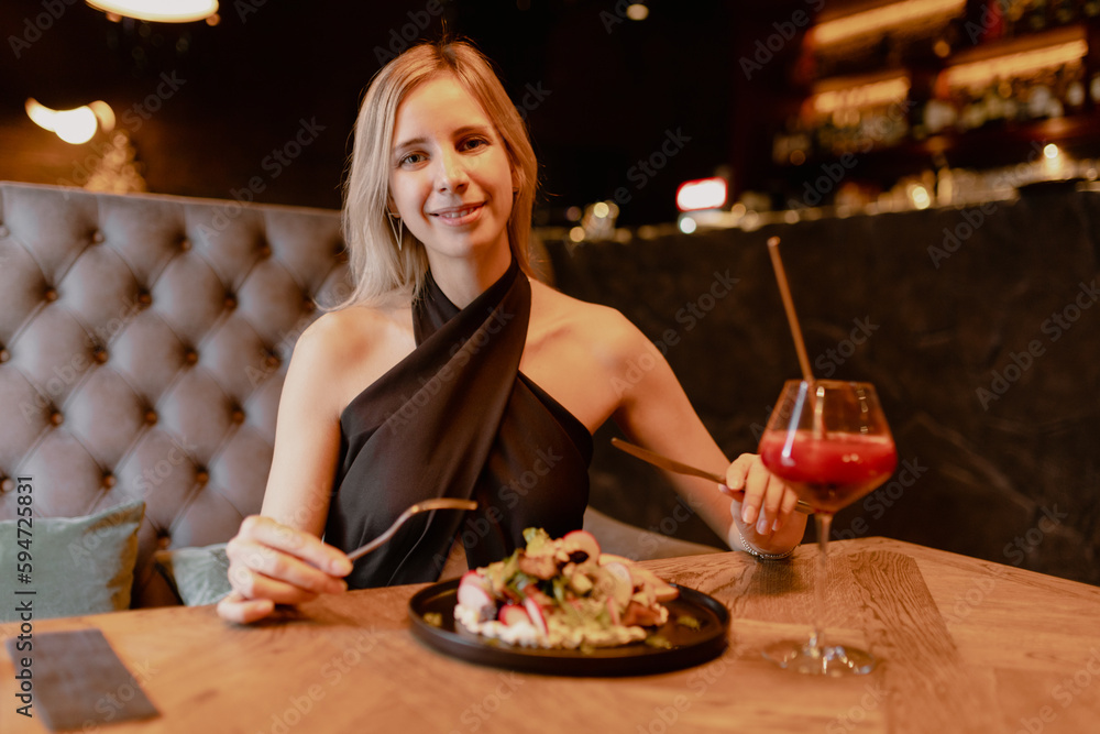 Portrait of young smiling attractive woman sitting on brown sofa at wooden table near black plate with vegetable salad and glass with red cocktail, holding fork, knife in restaurant cafe. Celebration.