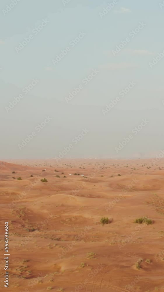 Drone view of a lifeless desert with sparse vegetation among red sand. Cars drive into a sandy mountain in the background Drone view of a lifeless desert with sparse vegetation among red sand. Cars drive into a sandy mountain in the background