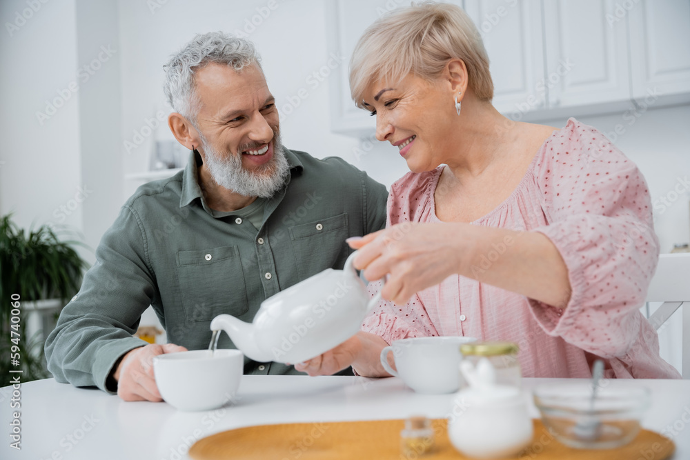 happy middle aged woman pouring tea near cheerful bearded husband in kitchen. happy middle aged woman pouring tea near cheerful bearded husband in kitchen.