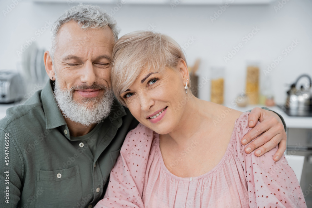 happy bearded man with closed eyes embracing wife smiling at camera in kitchen. happy bearded man with closed eyes embracing wife smiling at camera in kitchen.