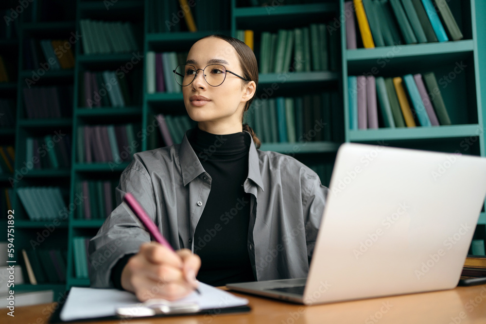 A woman sits at a desk with a laptop and a bookcase in the background. A woman sits at a desk with a laptop and a bookcase in the background.