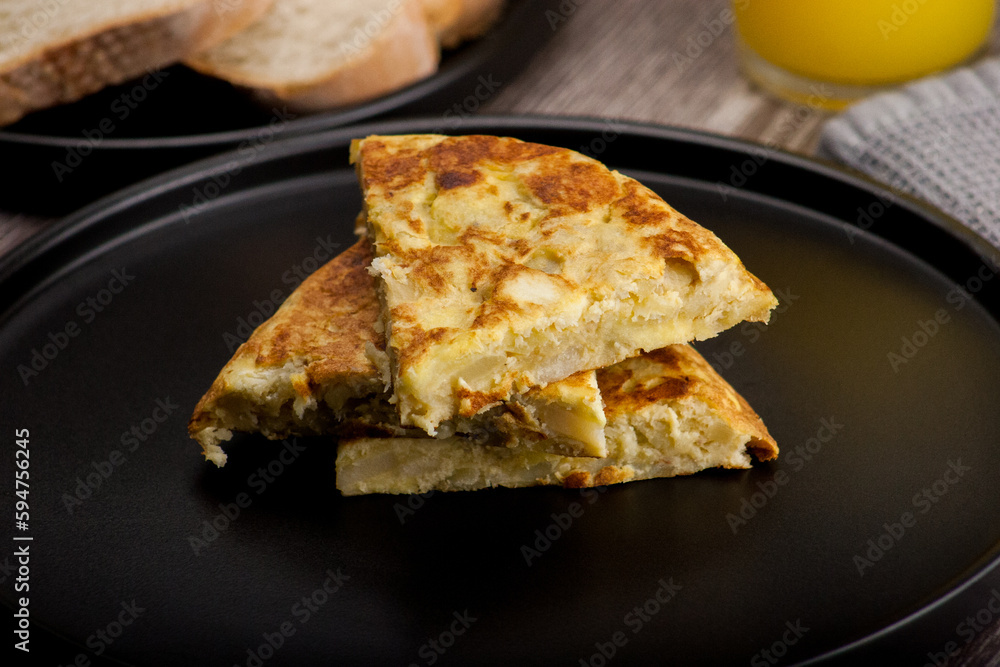 Traditional Spanish omelette with egg and onion served on a black plate, accompanied by a glass of orange juice and bread Traditional Spanish omelette with egg and onion served on a black plate, accompanied by a glass of orange juice and bread