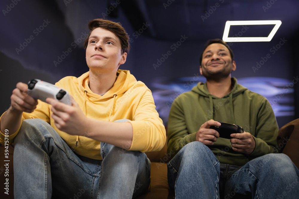 KYIV, UKRAINE - FEBRUARY 13, 2023: Low angle view of young man playing video game with blurred indian friend in gaming club. KYIV, UKRAINE - FEBRUARY 13, 2023: Low angle view of young man playing video game with blurred indian friend in gaming club.