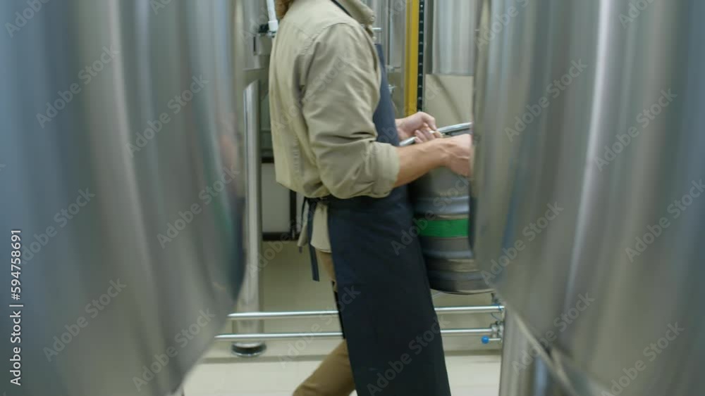 Lockdown slowmo shot of unrecognizable male craft brewery employee in apron carrying heavy keg of fresh lager beer or cider, filmed from behind large fermentation tanks Lockdown slowmo shot of unrecognizable male craft brewery employee in apron carrying heavy keg of fresh lager beer or cider, filmed from behind large fermentation tanks