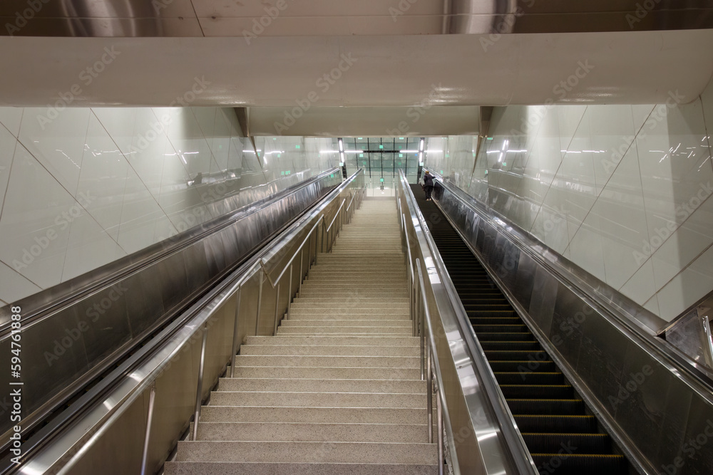 Looking up a long concrete stairs with an escalator on each side. A double door is at the top. White tile walls are on each side. Looking up a long concrete stairs with an escalator on each side. A double door is at the top. White tile walls are on each side.