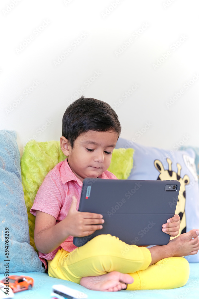 Little kid watching tablet gadget using touchpad. Happy small kid relax on couch in living room have fun browsing pad device online. Technology and education concept. Little kid watching tablet gadget using touchpad. Happy small kid relax on couch in living room have fun browsing pad device online. Technology and education concept.