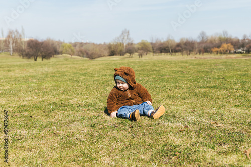 little toddler boy in a plush teddy bear hoodie sits on the grass in the park