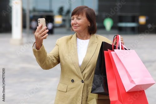 Stylish adult woman blogger with shopping bags and smartphone is communicating with followers in social networks near shopping mall. concept of consumerism, sale, rich life, virtual life. Black Friday