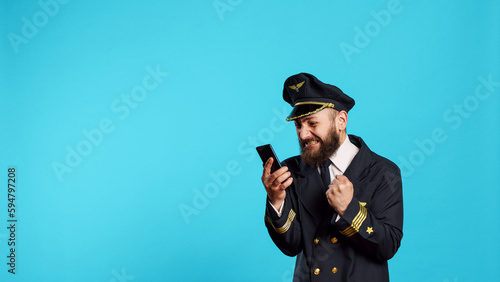 Young pilot reading good news on smartphone app, using onlien website and feeling happy about achievement. Male airline captain celebrating success on mobile phone, wearing uniform.