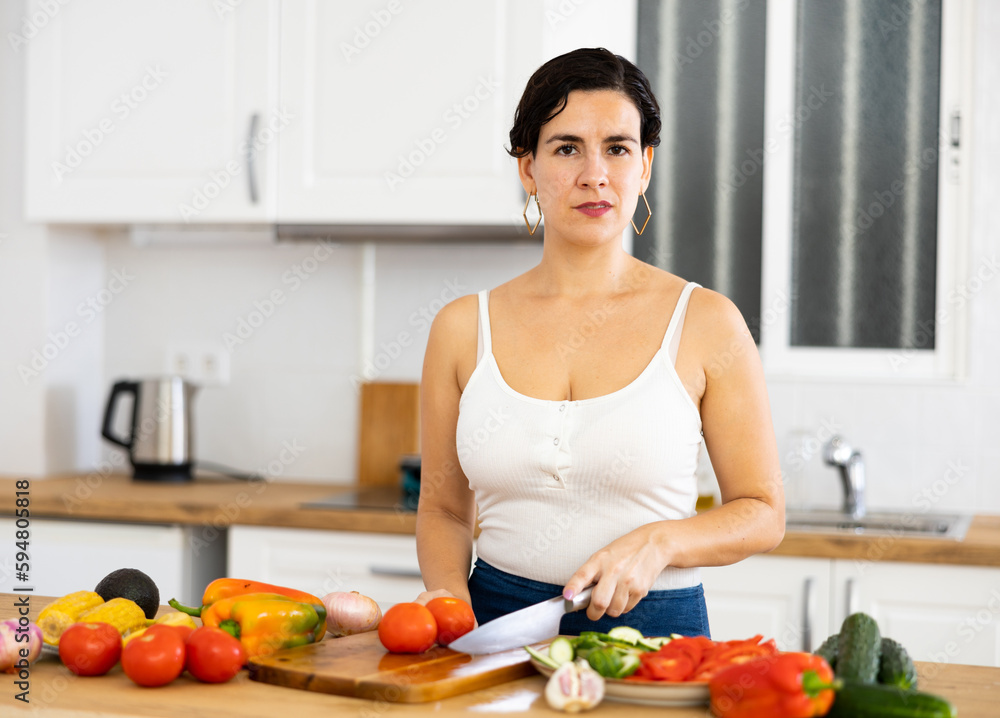 Smiling young Hispanic woman standing in home kitchen, preparing fresh vegetable salad. Vegetarian diet concept