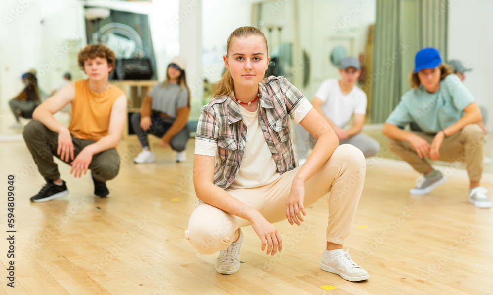 Confident teenage girl breakdancer posing during dance workout, squatting on haunches with group in choreographic studio