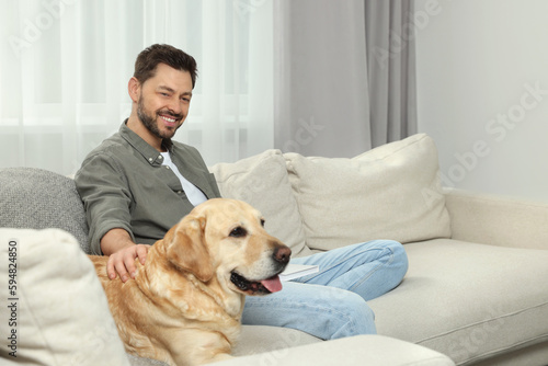 Man reading book on sofa near his cute Labrador Retriever at home. Space for text
