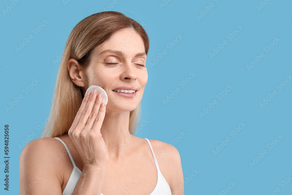 Fototapeta premium Beautiful woman removing makeup with cotton pad on light blue background, space for text