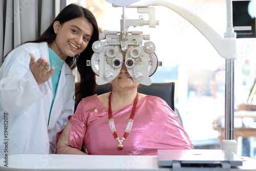 Indian or Nepali senior female customer looking through Optical Phoropter during eye exam, Pretty young optometrist taking care nearby, diagnostic ophthalmology equipment