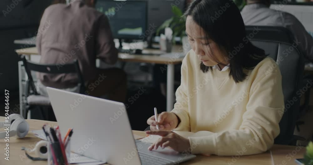 Young Asian woman analyst writing information in notebook and using laptop in open space office. Business and modern lifestyle concept. Young Asian woman analyst writing information in notebook and using laptop in open space office. Business and modern lifestyle concept.