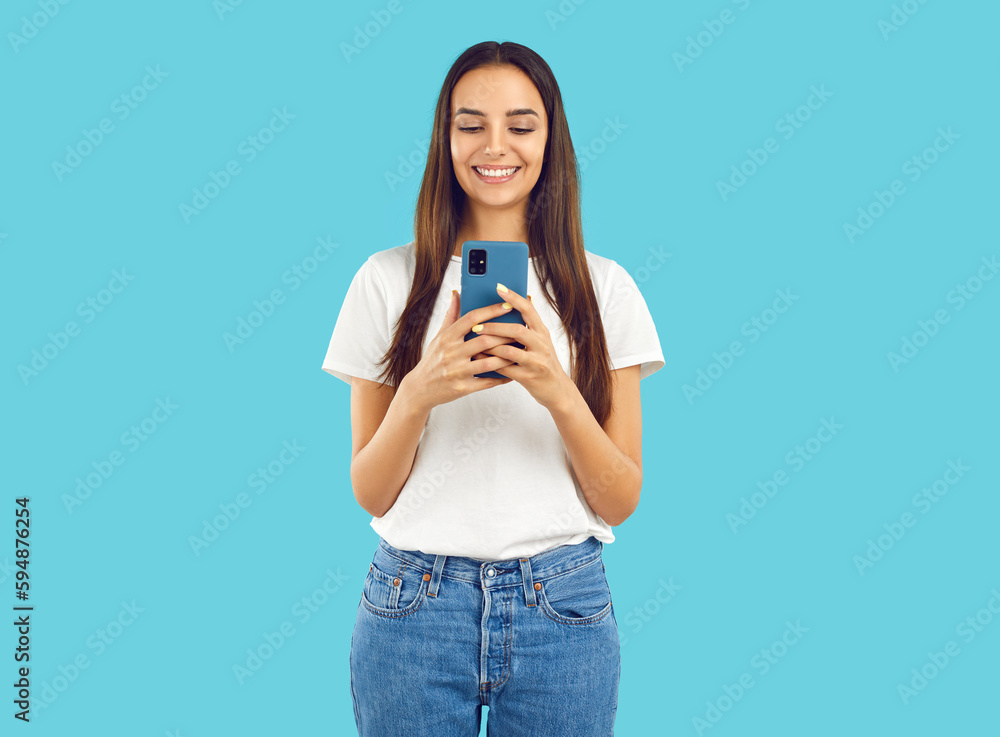 Happy smiling interested brunette girl with long hair in white t-shirt is looking at smartphone screen, reading message or watching video, talking video chat on blue background. Online life concept. Happy smiling interested brunette girl with long hair in white t-shirt is looking at smartphone screen, reading message or watching video, talking video chat on blue background. Online life concept.