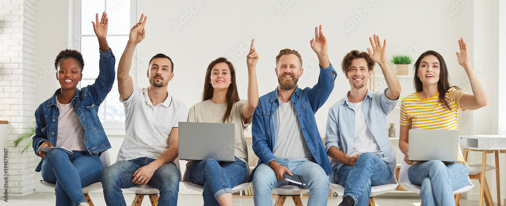 Group of satisfied multiracial listeners raise their hands to ask questions during training class. Young people in denim casual clothes sit in row on chairs with notebooks and laptops. Panoramic view. - obrazy, fototapety, plakaty 