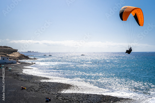 Unrecognizable paraglider lands on the Playa de la Enramada beach, a renowned paragliding spot in Tenerife, as the sparkling Atlantic Ocean serves as a picturesque backdrop, illuminated by sunlight. photo