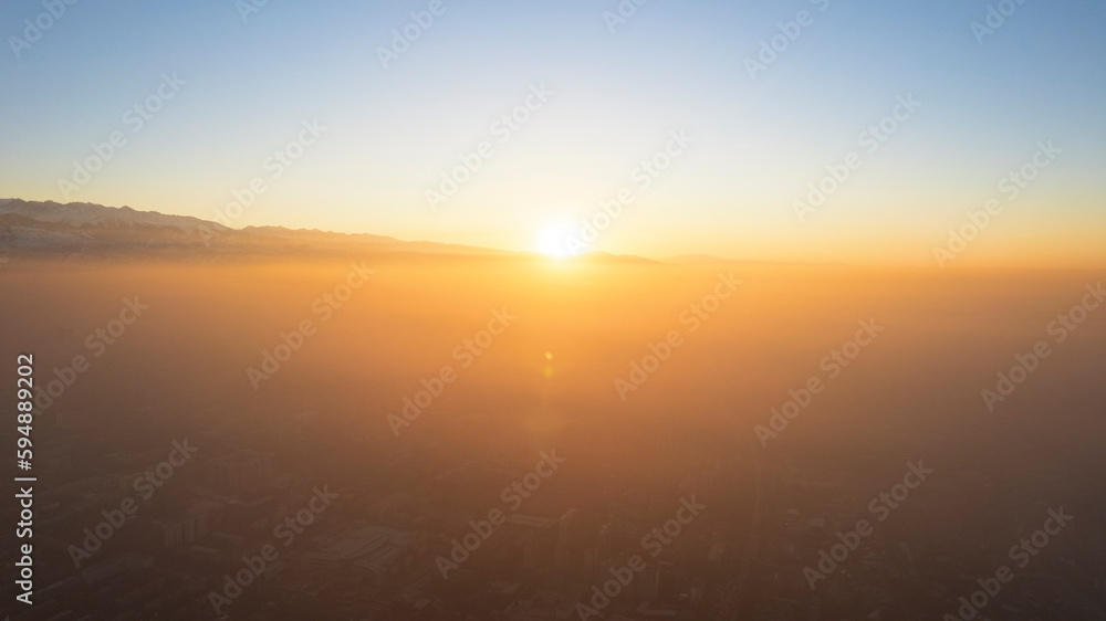 Epic gray smog is visible at sunset over the city. A bird's-eye view from a drone of houses, roads, cars and parks. White clouds and snowy mountains are illuminated by orange rays of the sun. Almaty