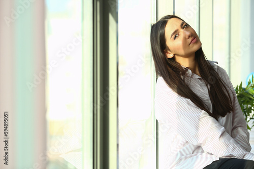 Dreamy young business woman is sitting on windowsill near huge window in modern office sun is shining brightly. Businesswoman enjoying break and relaxing after a hard day at work