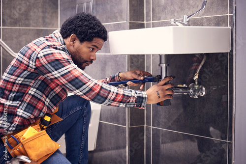 Black man, plumber and maintenance in bathroom, fixing sink pipe with wrench and plumbing industry. Manual labor, male with focus and trade, handyman doing repairs with tools on home renovation