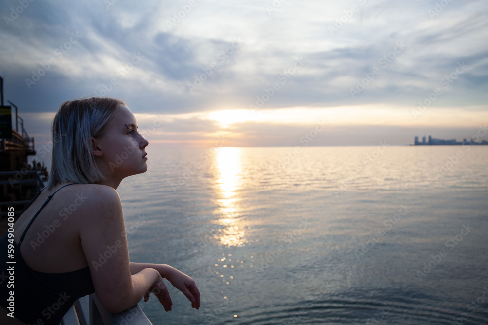 Obraz premium Beautiful blonde woman in tank top standing on pier on the background of city silhouette, cloudy sky and sea