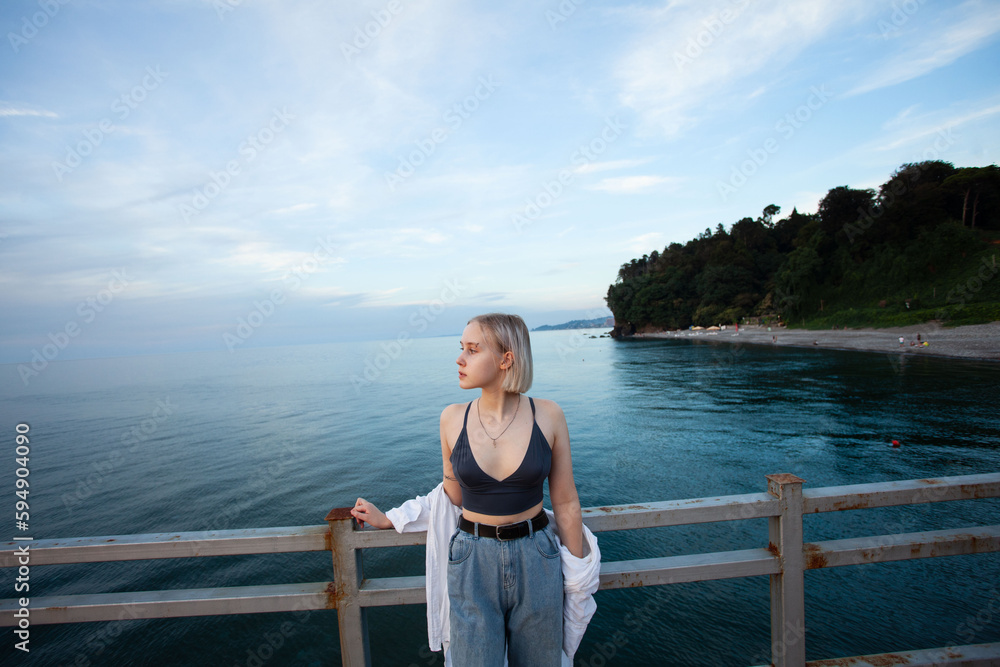 Cute stylish blonde woman posing on pier on the background of sea, cloudy sky and coastline with green trees