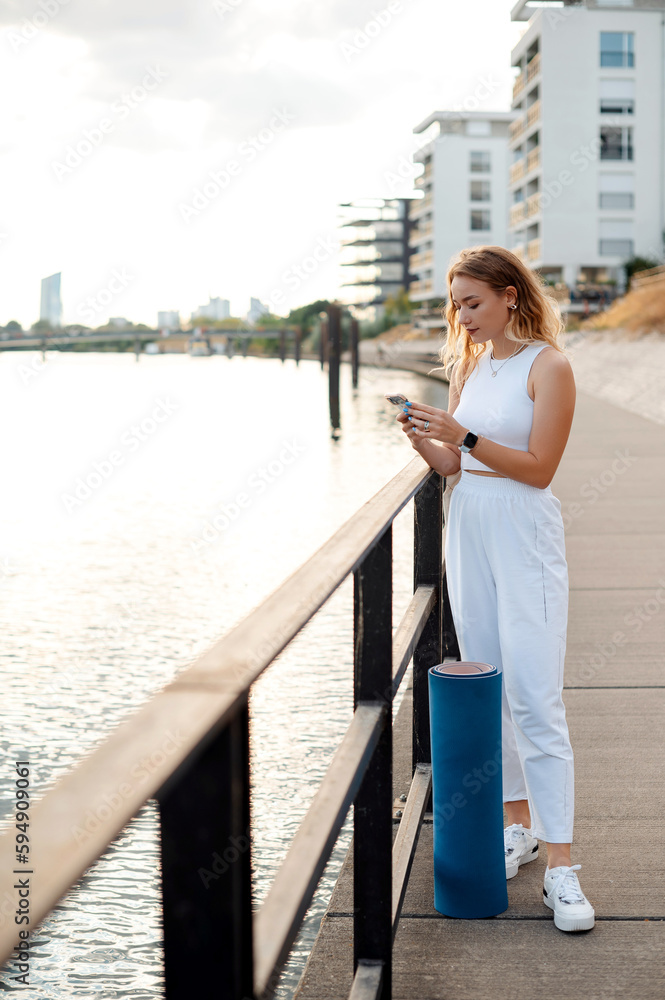 Vertical view of the young lady standing at the river embankment and chatting smartphone