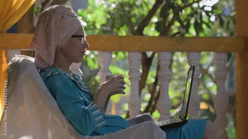 Caucasian woman using her laptop while sitting on the balcony after a shower in a bathrobe with a towel on her head. Female freelancer communicates via video link on a terrace in sunlight. Caucasian woman using her laptop while sitting on the balcony after a shower in a bathrobe with a towel on her head. Female freelancer communicates via video link on a terrace in sunlight.