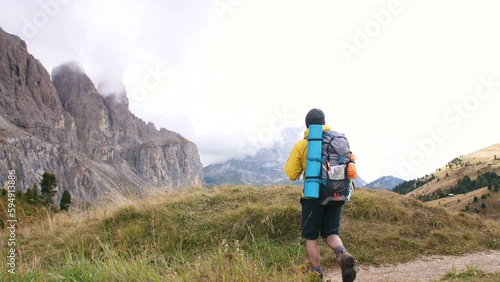 Arrival. Young man hiker hiking on mountain dirt road with cloudy sky and fog. Yellow jacket, backpack, black beard and beanie. Traveling Dolomites, Italy.