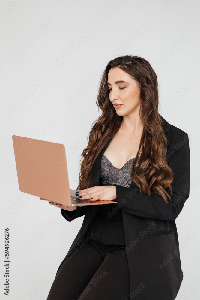 young girl with long dark curly hair in a black suit with a laptop sits on a white background. Businesswoman working online young girl with long dark curly hair in a black suit with a laptop sits on a white background. Businesswoman working online