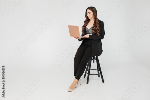 young girl with long dark curly hair in a black suit with a laptop sits on a white background. Businesswoman working online