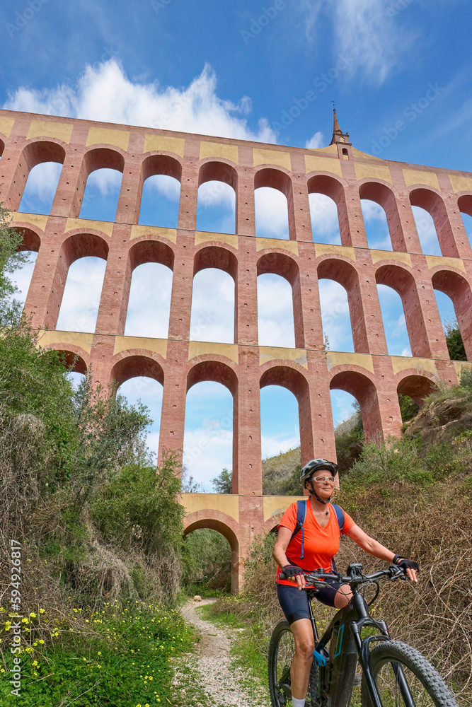 nice senior woman cycling with her electric mountain bike below the aqueduct of Aguila in the Sierra de Tejada near Nerja, Andalusia, Spain
