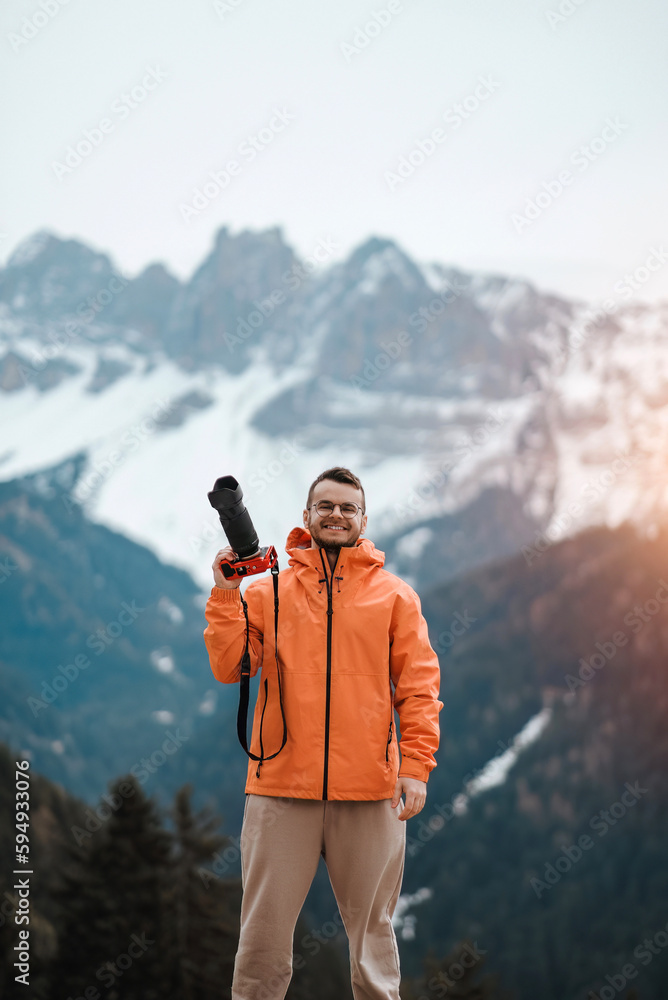 The young man holds the mirrorless camera in his hand while the alpine mountains rise majestically in the background and their snow-capped peaks reaching towards the sky.
