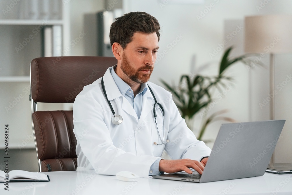 By the table with laptop. Doctor in formal clothes is working in the cabinet By the table with laptop. Doctor in formal clothes is working in the cabinet