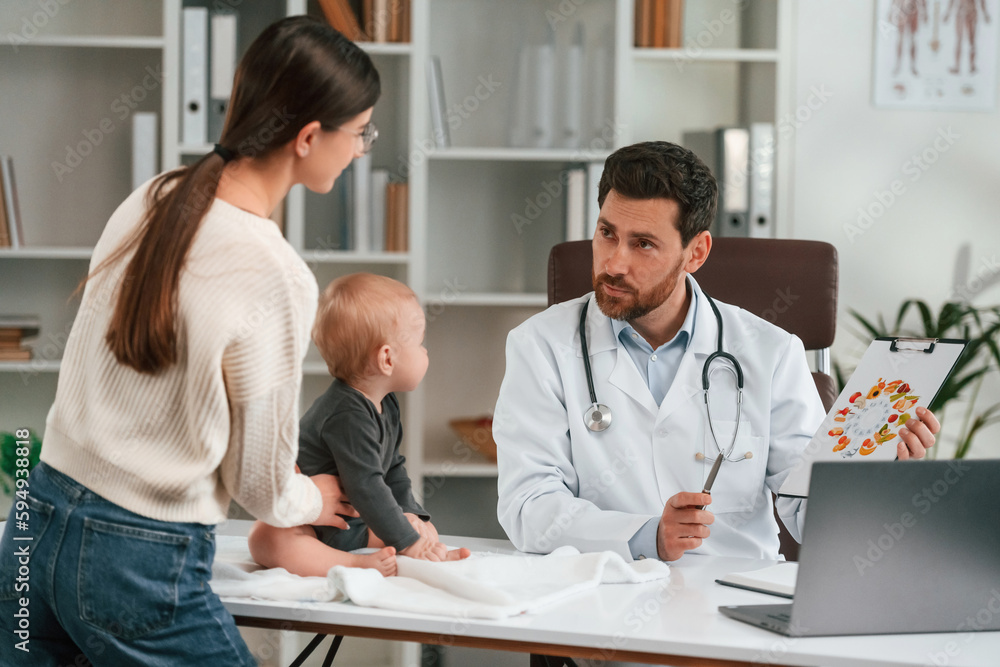 Fototapeta premium Having consultation. With baby and mother. Doctor in formal clothes is working in the cabinet