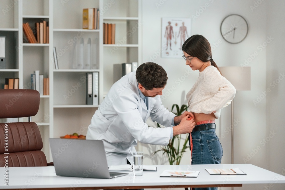 Fototapeta premium Measuring the waist. With woman. Doctor in formal clothes is working in the cabinet