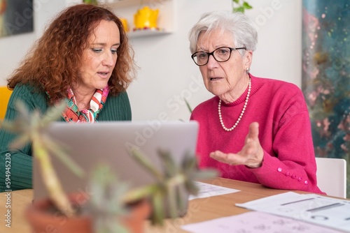 Redhead woman sitting at table with gray haired mother in glasses having confused face expression while using computer at home