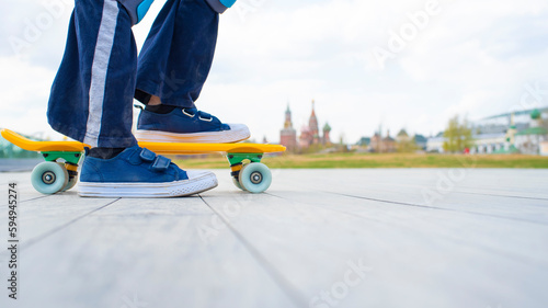 feet of a child in blue sneakers on a yellow plastic city cruiser, sketord on the road in the Zaryadye park. in the distance the Kremlin, red square, St. Basil's Cathedral in Moscow. spring, summer.