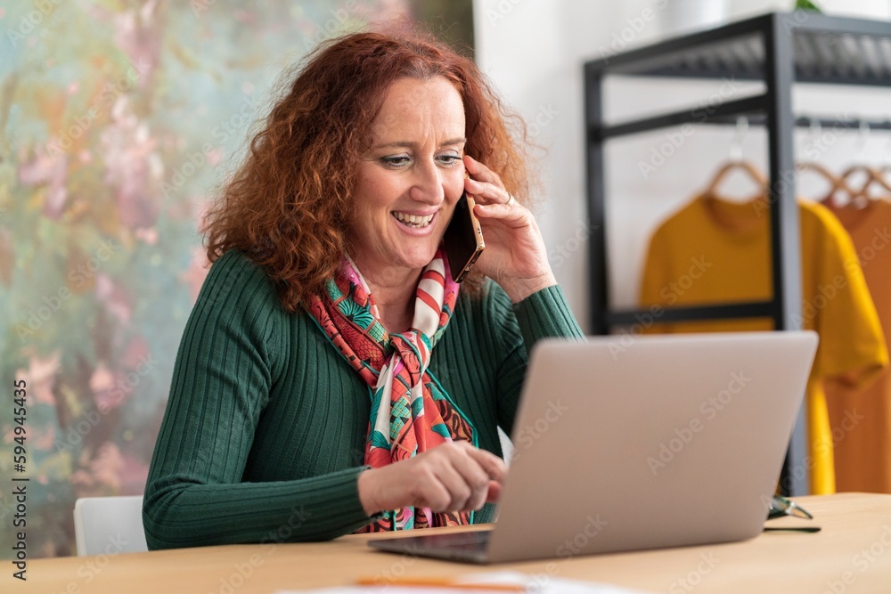 Mature redhead haired woman in green sweater sitting at table having conversation via mobile phone while browsing computer Mature redhead haired woman in green sweater sitting at table having conversation via mobile phone while browsing computer