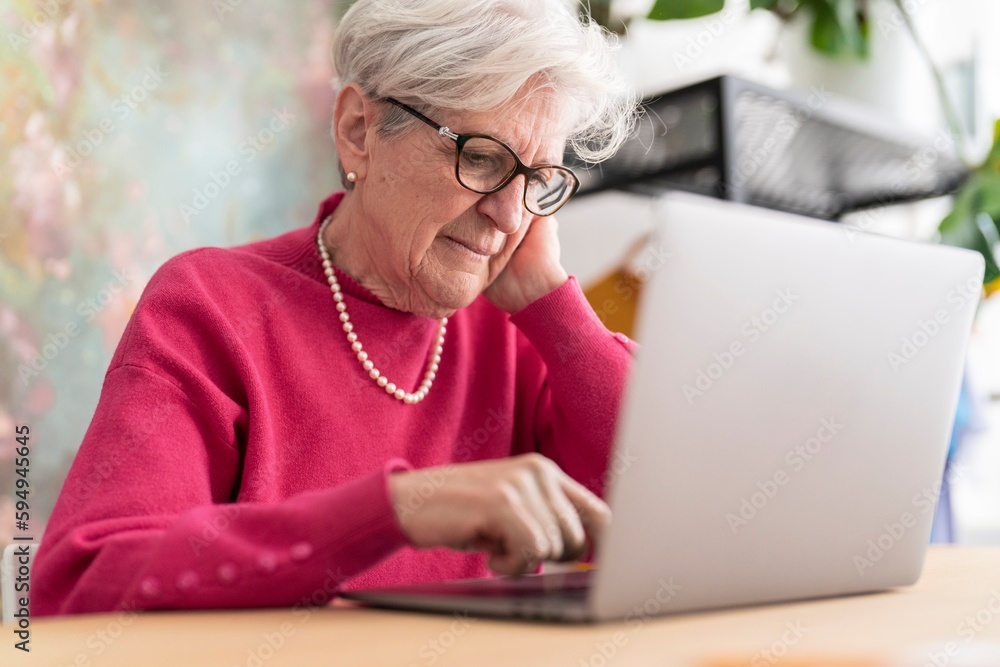 Positive elderly lady in glasses and pink sweater sitting at table browsing computer surfing internet at home Positive elderly lady in glasses and pink sweater sitting at table browsing computer surfing internet at home