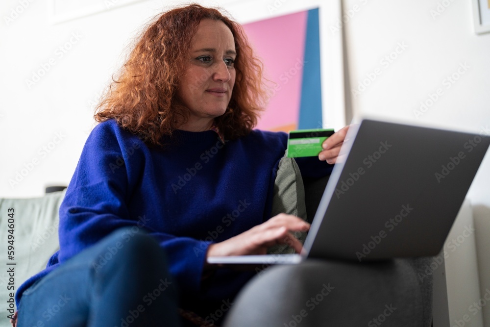 Positive middle aged redhead female in blue hoodie sitting on sofa and using computer while paying online for purchases with credit card Positive middle aged redhead female in blue hoodie sitting on sofa and using computer while paying online for purchases with credit card